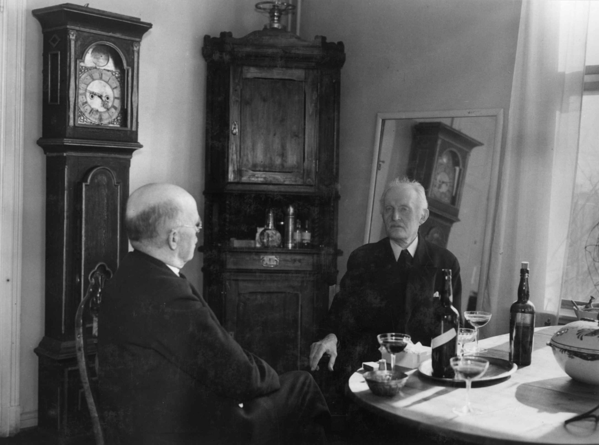 Kunstneren og maleren Edvard Munch fotografert i 1937 på Ekely.  Sitter ved spisebordet i samtale med fotograf Anders Beer Wilse. Flasker og tre glass til servering på bordet. Interiør med klokke og hjørneskap ved vindu.