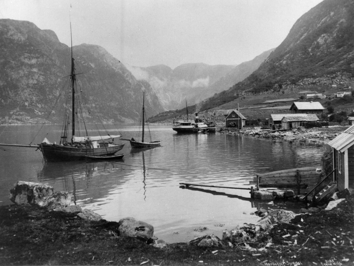 Seilbåt og dampskip på fjorden i Sundal i Mauranger i Kvinnherad i Hordaland. Naust, brygge og fjell. Fotografert i perioden 1885 - 1892.