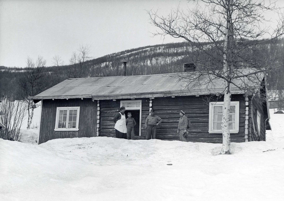 En kvinne, en gutt og to menn står utenfor en hytte eller et seterhus i laftet tømmer på Geilo. Snødekt vinterlandskap.