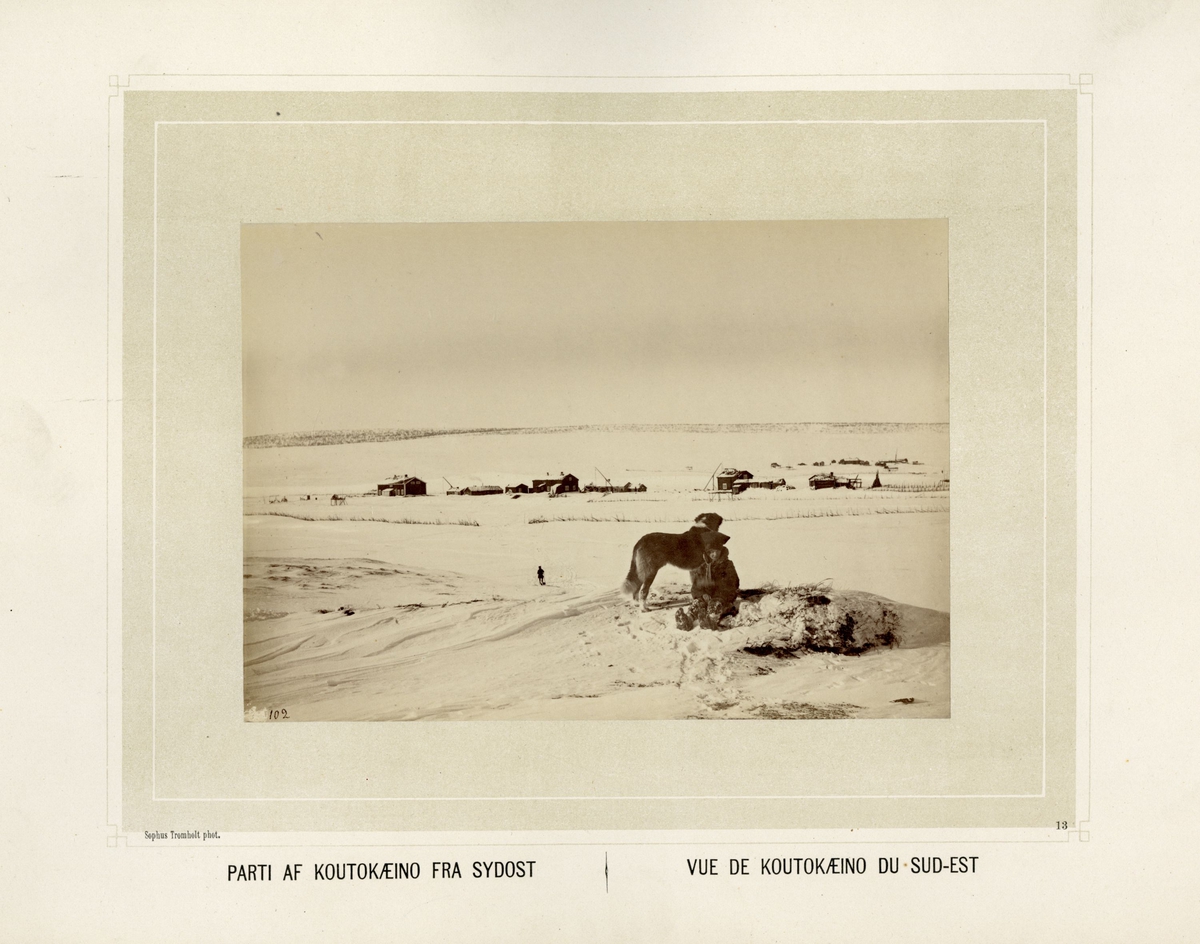 Kautokeino sett fra sydøst med bebyggelse. Person i samisk drakt sitter i forgrunnen sammen med en hund.