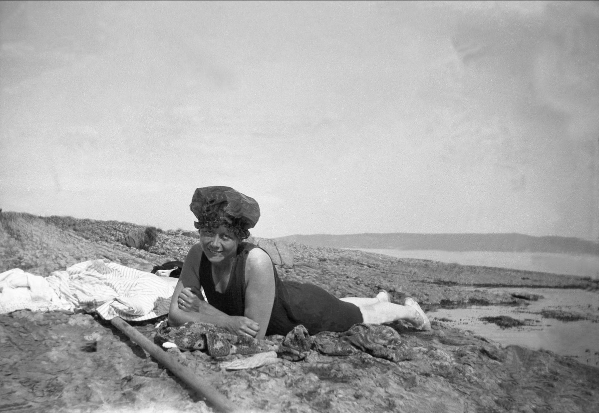 En kvinne med badedrakt ligger på magen å poserer på en strand. Robsahm og Lund.