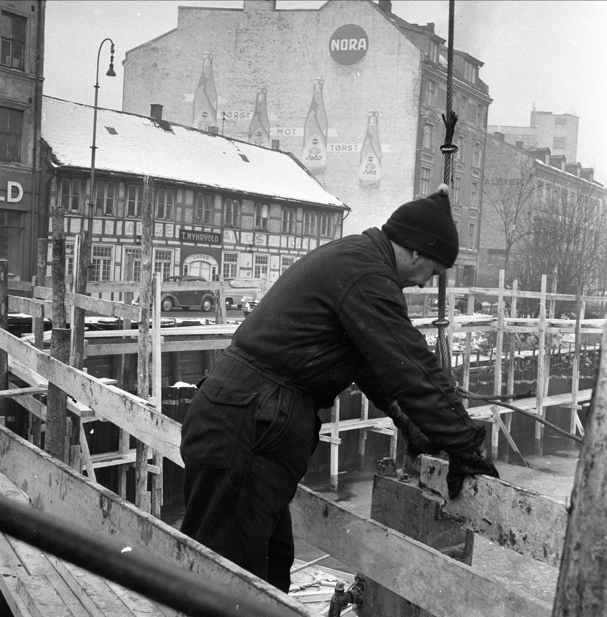 Schweigaards bru, Oslo, 23.02.1956. Anleggsarbeid. Bebyggelse og elva, mann på stilaser. Bindingsverksbygning i Rødfyllgata 12 før riving, nå på Norsk Folkemuseum.