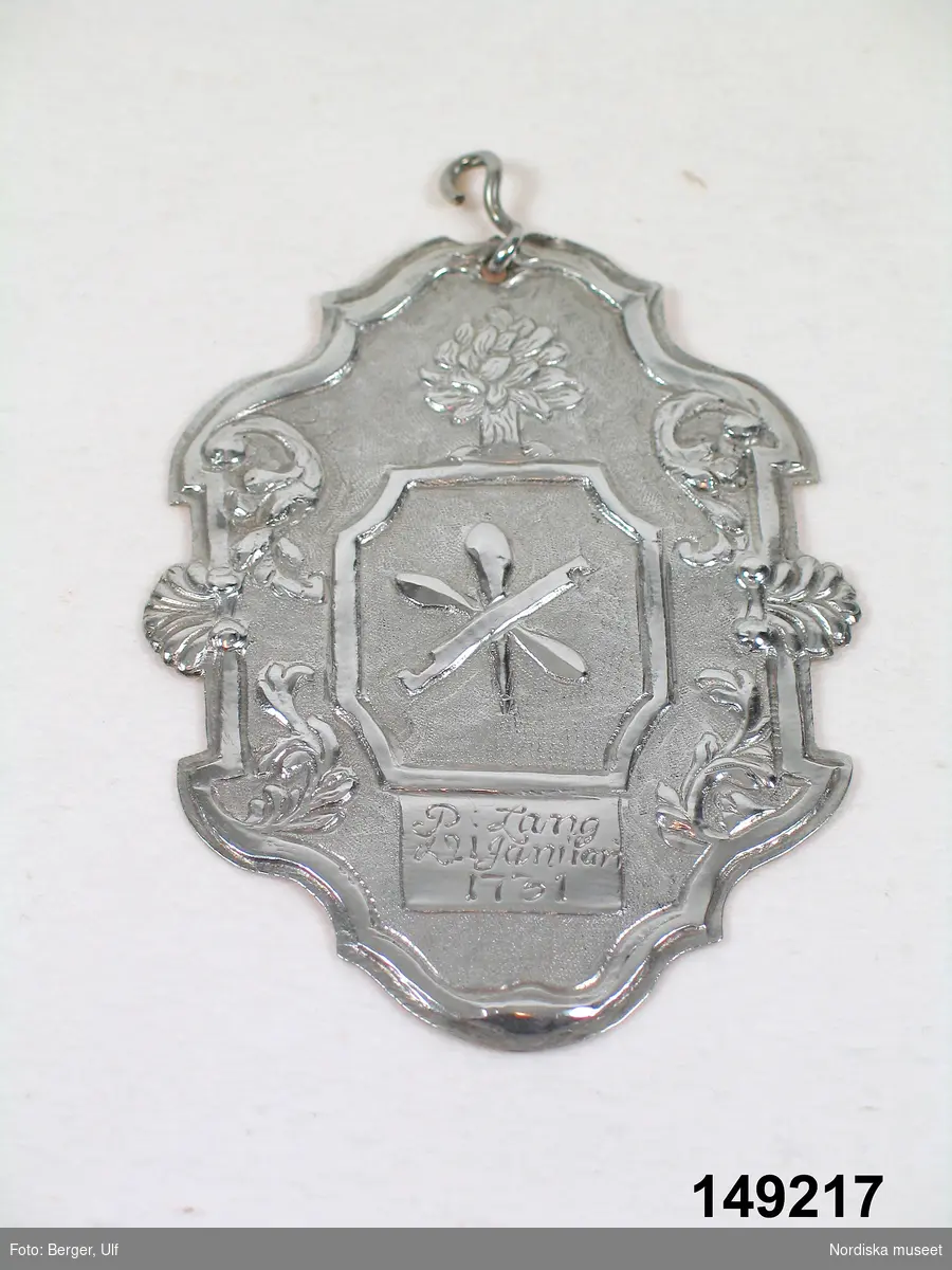 Huvudliggaren:
"Skråskylt, tillhörande glasmästargesällskapets välkomma (inv.nr 149.142), af silfver, drifven ornering med glasmässtare emblem. Inskr. P. Lang d 21 Januari 1731'."