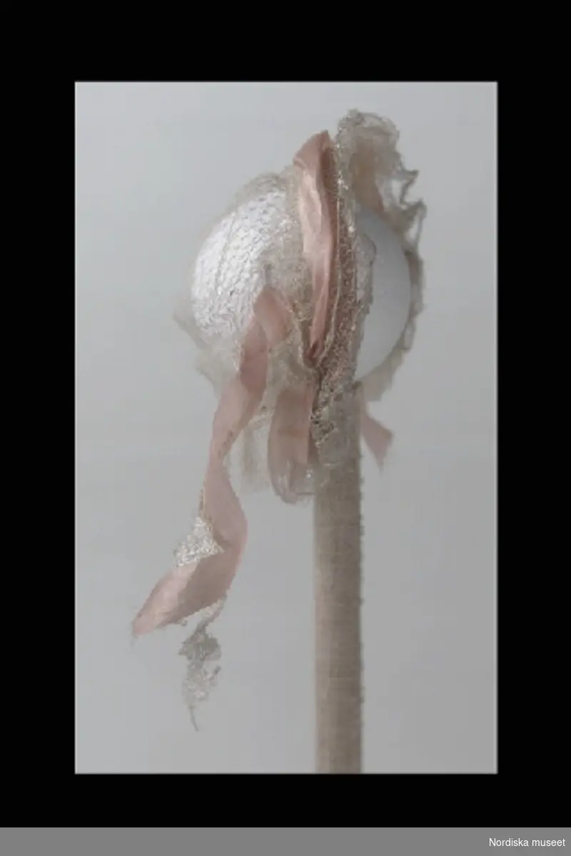 Inventering Sesam 1996-1999:
H 7 cm
Dockhårklädsel av vit tyll, dekorerad runt överkanten med rosa sidenband, metalltråd runt kanten.
Hör till docka 202.174.
Leif Wallin jan 1997