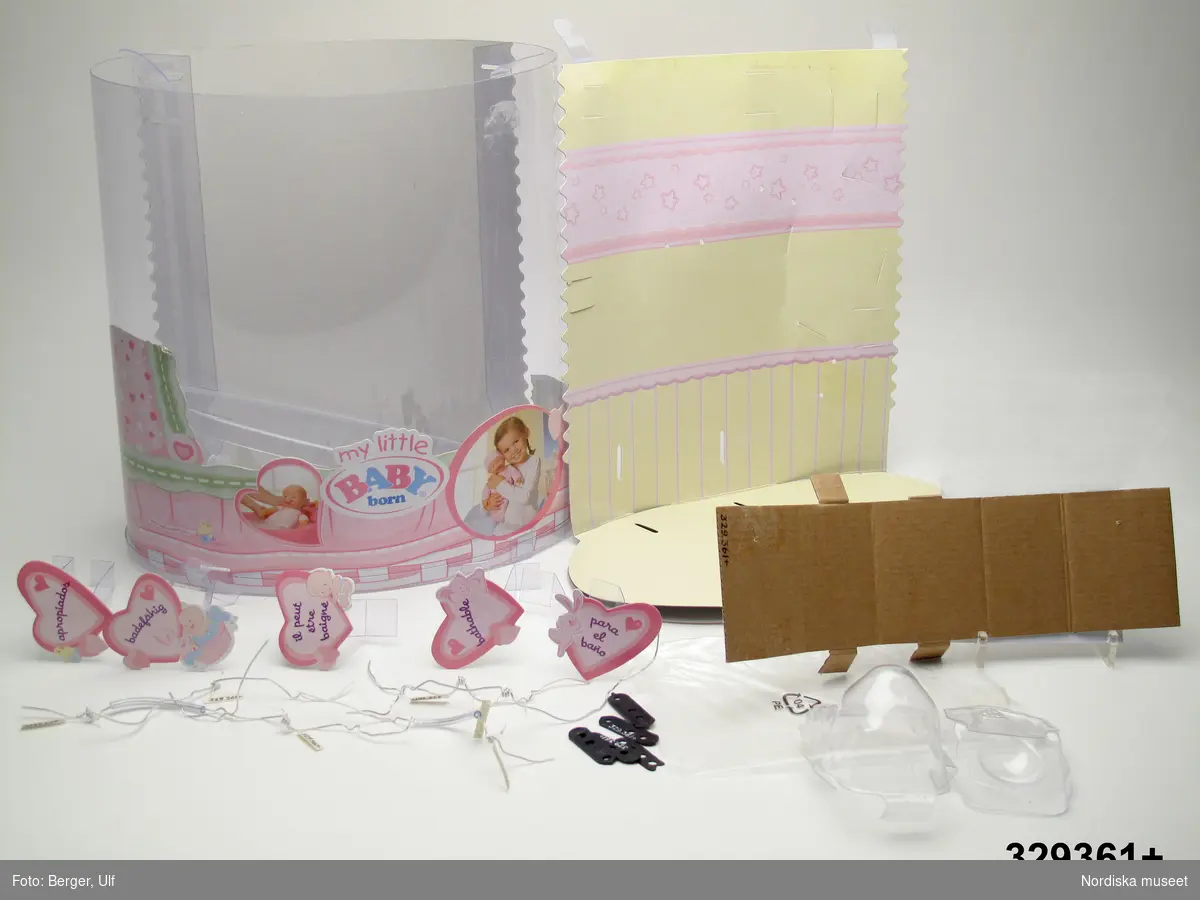 Förpackning till dockan "My little Baby born" i genomskinlig plast och ellipsformat tvärsnitt. Längd: 30,5 cm, bredd: 18 cm, höjd: 31 cm.
I förpackningen konstruktioner för förvaring av dockan med tillbehör. På fram- och baksida utanpå plasten på papp och i färgtryck information och diverse instruktionsbilder om föremålet, upplysning om att leksaken är lämplig för barn från ett år och uppåt, varumärke mm. 
Upptill och nertill på förpackningen avmonteringsbara ellipsformade lock i genomskinlig plast. 
Inuti förpackningen en pappdel i gult och rosa som bildar baksida och golv. På undersidan (ena elipsformade golvdelen) finns information på olika språk. 
5 st hängande hjärtan i papp på genomskinliga plastdelar (som fästes i pappväggen och på baksidan var fastsatta med genomskinlig 4,8 cm bred tejp), med information på olika språk om att dockan är möjlig att bada. 
Genomskinlig plastficka för informationsfolder. 6 st plastklädda ståltrådar (varav två med ytterligare plastförstärkt mittdel) som skall hålla dockan på plats i förpackningen. 5 st svarta förstärkningsdelar i plast med tre genomgående hål i vardera. En är skadad i ena kortänden. (Dessa har suttit på baksidan av pappförpackningen där ståltrådarna kommit ut och slutits till). 
Vit kartongdel som bildat en kub i botten på förpackningen. Två genomskinliga plastdelar som varit förpackning åt skallran.

Se tillhörande docka inv.nr 329360a-f.

Rekonstruktion av julklappsönskemål julen 2007 från Minna Skantz, född 1999, Vaxholm. Ingår i projektet "Barn tar plats". Se dokumentation i arkivet D455.
/Ulf Hamilton 2008-05-29      