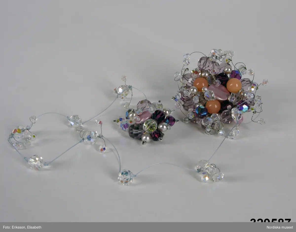 Dräktsmycke i form av två broscher, en större och en mindre, tillverkade av glasstenar i vitt, rosa, violett och aprikos, uppträdda på silverfärgad metalltråd och fästade oregelbundet mot mässingsplatta med nål och krylle. De två broscherna är sammanlänkade med nylontråd med påfästade glasstenar. 

Använd  på rosa galaklänning (inv.nr 329.407a-b) med capeliknade schal till. Smycket var fästat med större broschen på schalen och den mindre på kjoldelen i höfthöjd. Allt arrangerat av Gunilla Pontén, klänningens formgivare. 

Komponerad av Gunilla Pontén till Karin Hyldgaard -Jensen, vinnare av Fröken Sverige-titeln 1962. Vinnaren tävlade senare under året om titeln "Miss International Beauty" i Long Beach, Californien. Där kom Karin med till semifinalen. Hela resegarderoben komponerades av Gunilla Pontén. Den kompletterades med konfektionsplagg som också valdes ut av G.P. Till detta kom också en "folkdräkt" som var tung och varm och inte särskilt populär hos Karin.
Se inv.nr 329.405-329.407 samt tillbehör 329.585-329.587
/Ingrid Roos 2009-06-04
