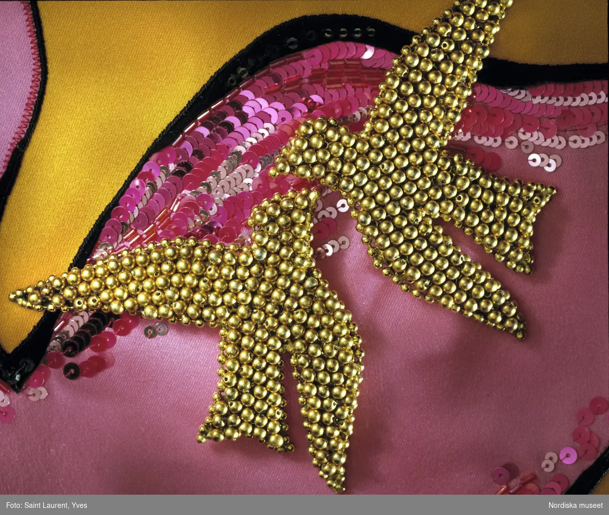 Hellång aftonklänning med axelbandslöst liv av sidenduchesse. Livet ceriserosa, kjolen orangegul med applikationer i form av stiliserade fåglar i rosa siden prydda med paljetter och pärlbroderi. Från Yves Saint Laurents vårkollektion 1988. Nordiska museet inv nr 318887.