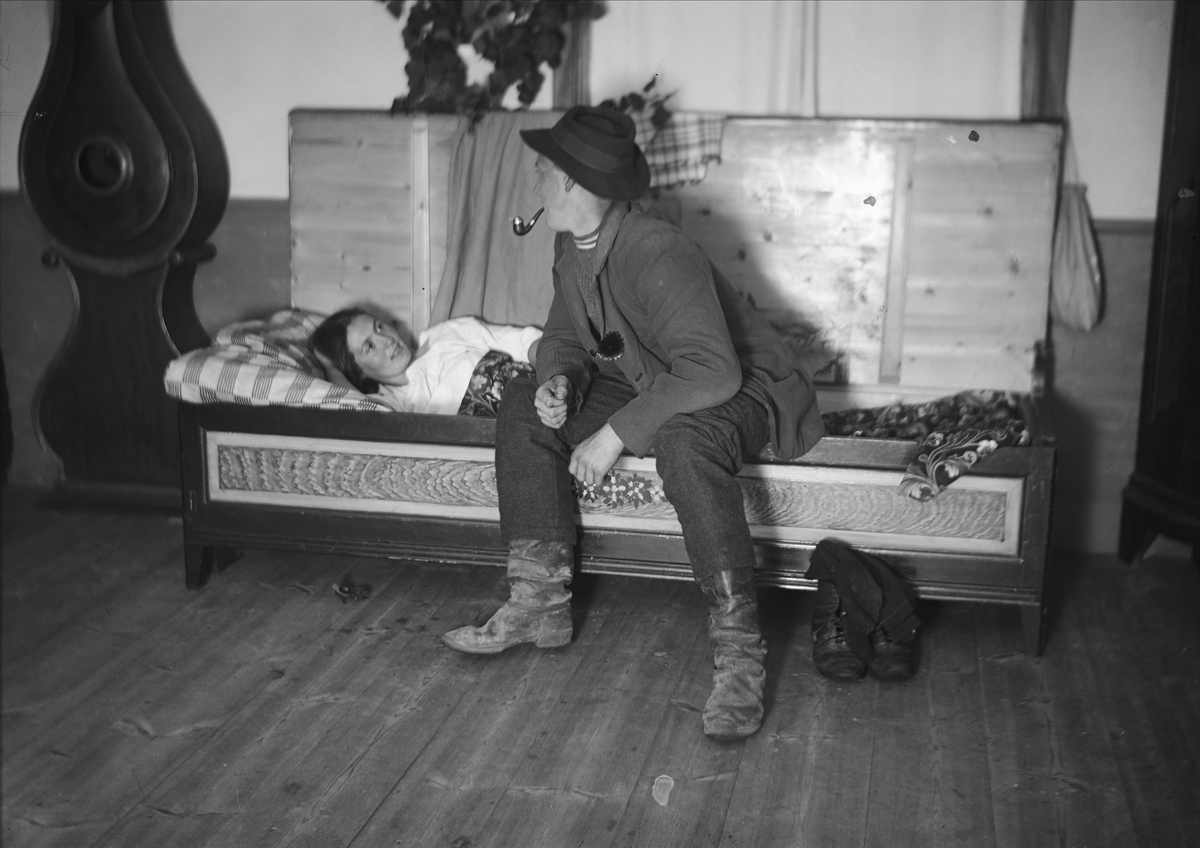 Nattfrieri. En man sitter på sängkanten och pratar med liggande kvinna. Värmland, Mangskogs socken.