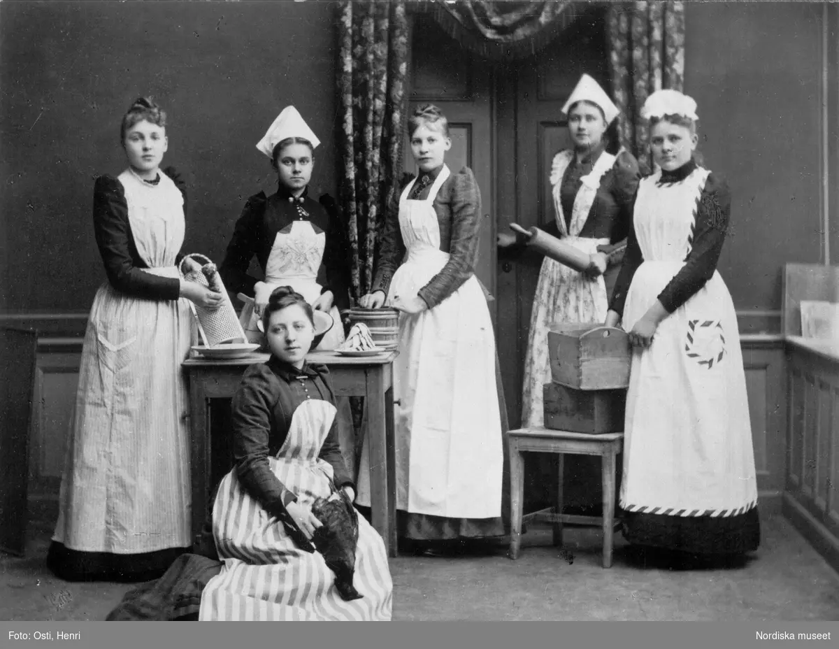 Elever vid Uppsala Hushållsskola. Flickorna är klädda i mörka klänningar och ljusa förkläden, tre stycken har en vit hätta på huvudet. Flickorna håller i olika hushållsredskap. Bakom skymtar en dörr och ett draperi.