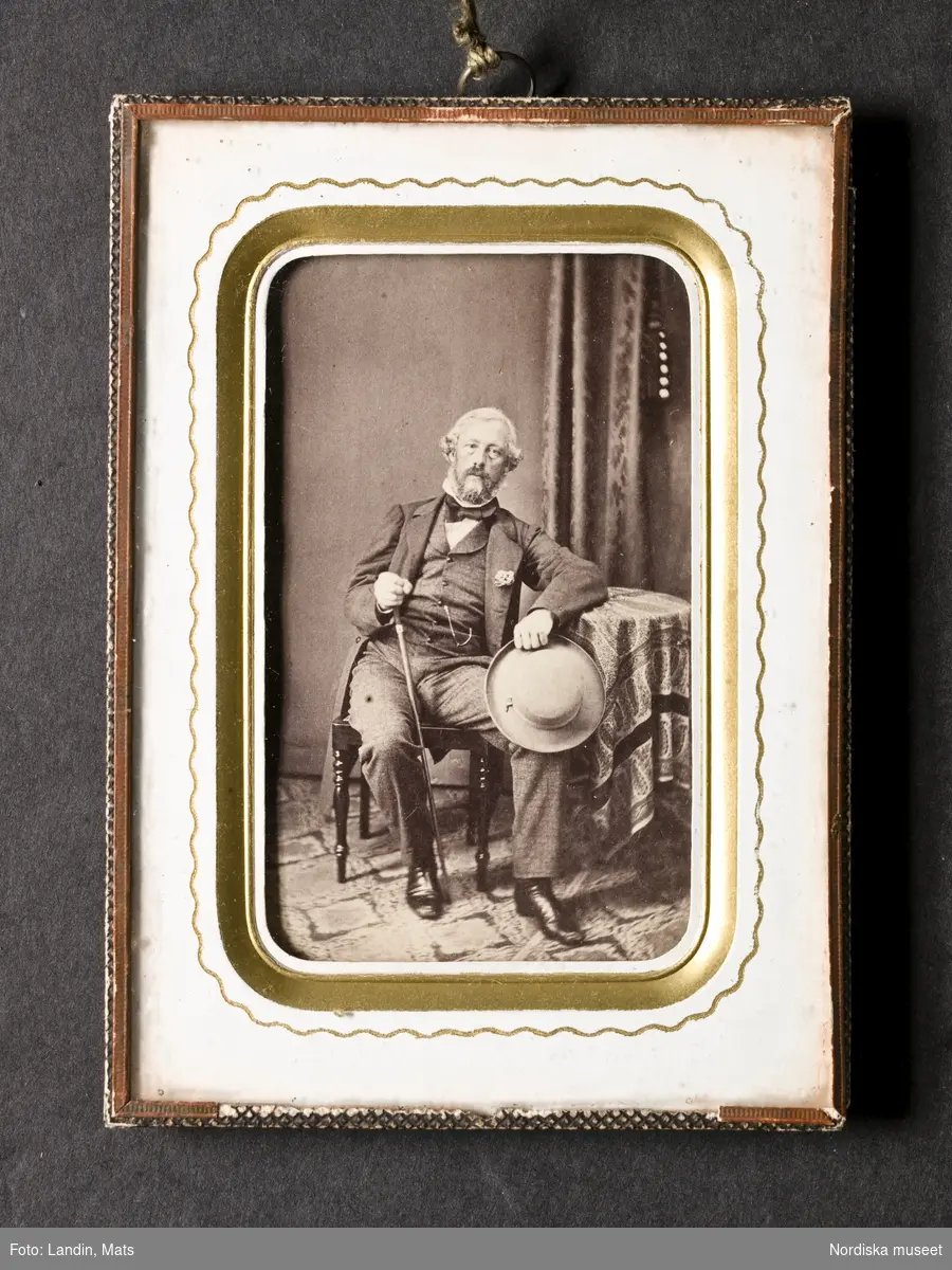 Porträtt av okänd man, sittande vid ett bord med hatt o käpp i händerna. Med skägg och mustasch, klädd i bonjour, rutiga byxor, väst, vit skjorta och svart halsduk. Fotografi i ram, porträtt i visitkortsformat.
Nordiska museet inv.nr 185541.