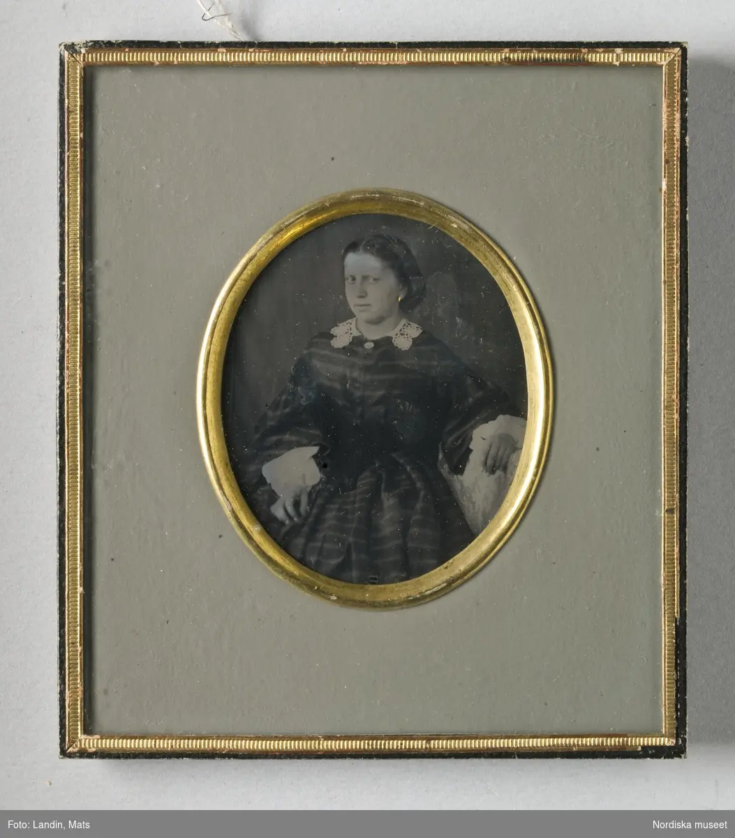 Porträtt, knäbild, av sittande ung kvinna med mittbenat hår, mörk klänning, vita manschetter och vit virkad krage. Förgyllt örhänge. Ambrotypi i ram, 
Nordiska museet inv.nr 249995.
