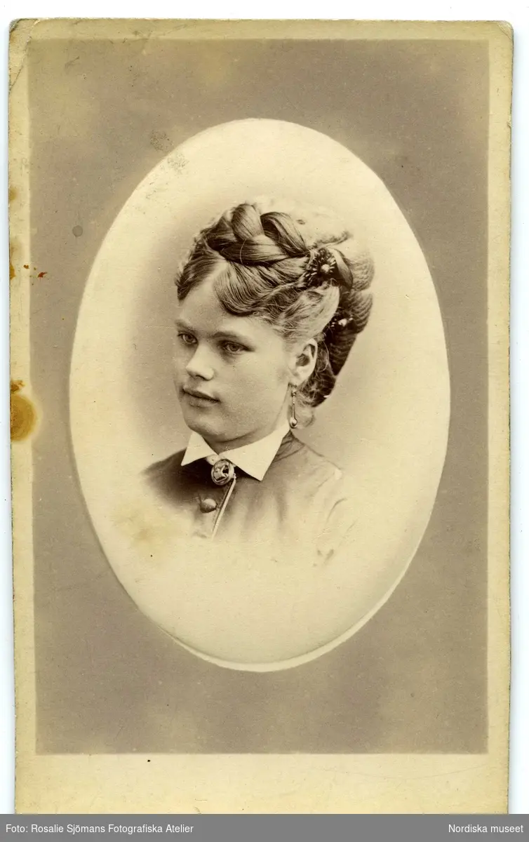 Ung kvinna med konstfull frisyr med blomformade hårsmycken, örhängen och brosch. 1870-tal.
