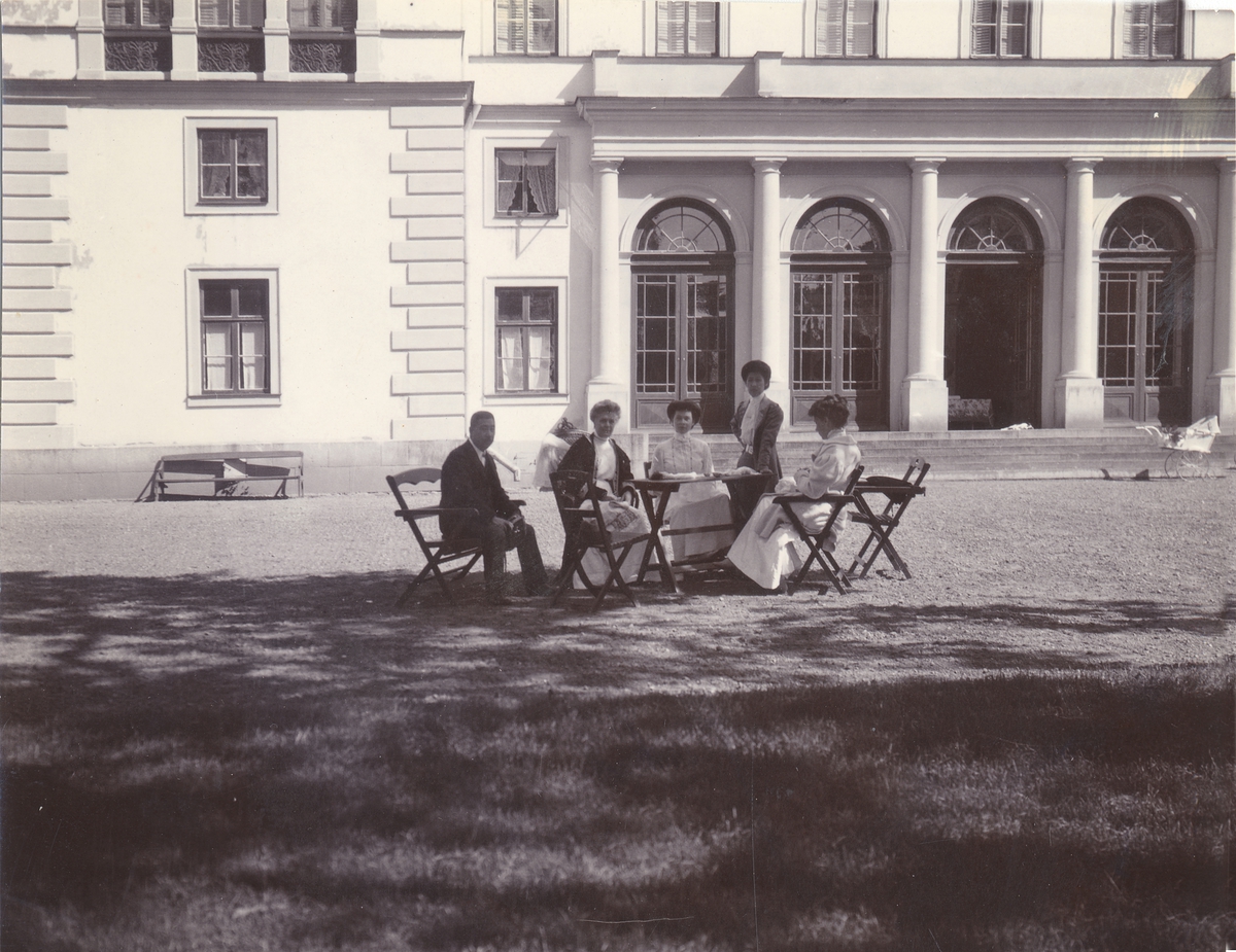 Ett sällskap sitter runt ett bord på gården framför en slottsbyggnad.