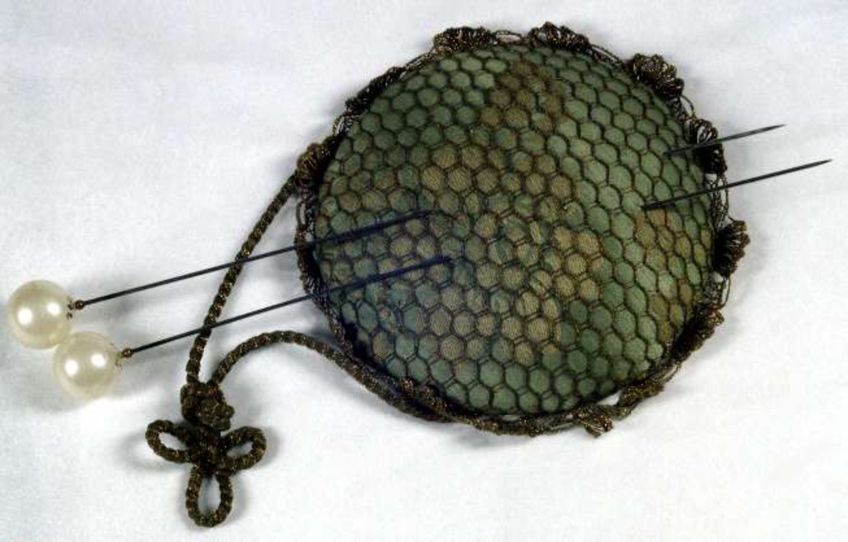 Dyna klädd med ljust blågrönt sidentyg överklädd med nät av gulmetalltråd. Spetskant av samma material. Två hattnålar UM07792 istuckna i dynan.