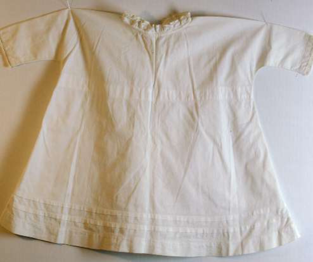 Barnklänning av vitt tuskaftat bomullstyg med invävda prickar, stråveck ovanför fållen, brodyr runt ärmar och halsringning. Knyts med bomullsband i dragsko i halsen och knäpps med fyra knappar i ryggen. Maskinsydd.
