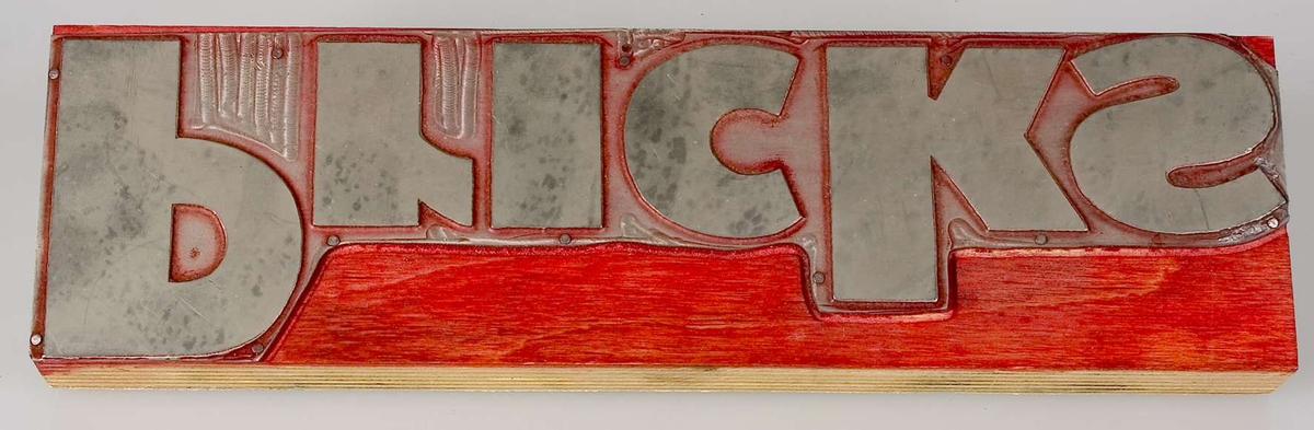 En kliché av metall, monterad på en rektangulär platta av skiktlimmat trä. Rester av färg. Text: Pricks.