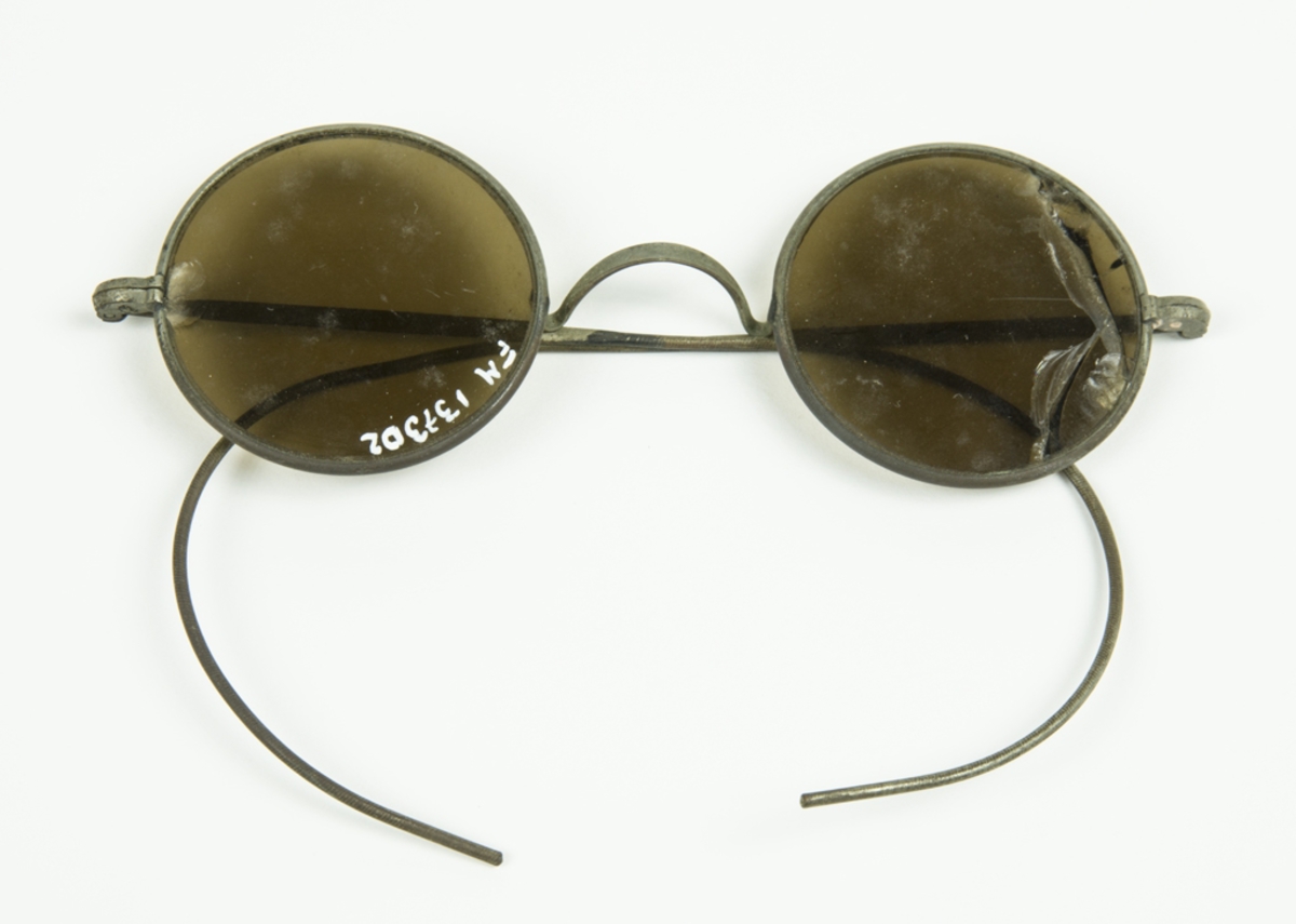 Solglasögon med metallskalmar med små runda glas. Ena glasögat är sprucket och limmat. Öronbågen går runt hela örat (typ idrottsvariant).