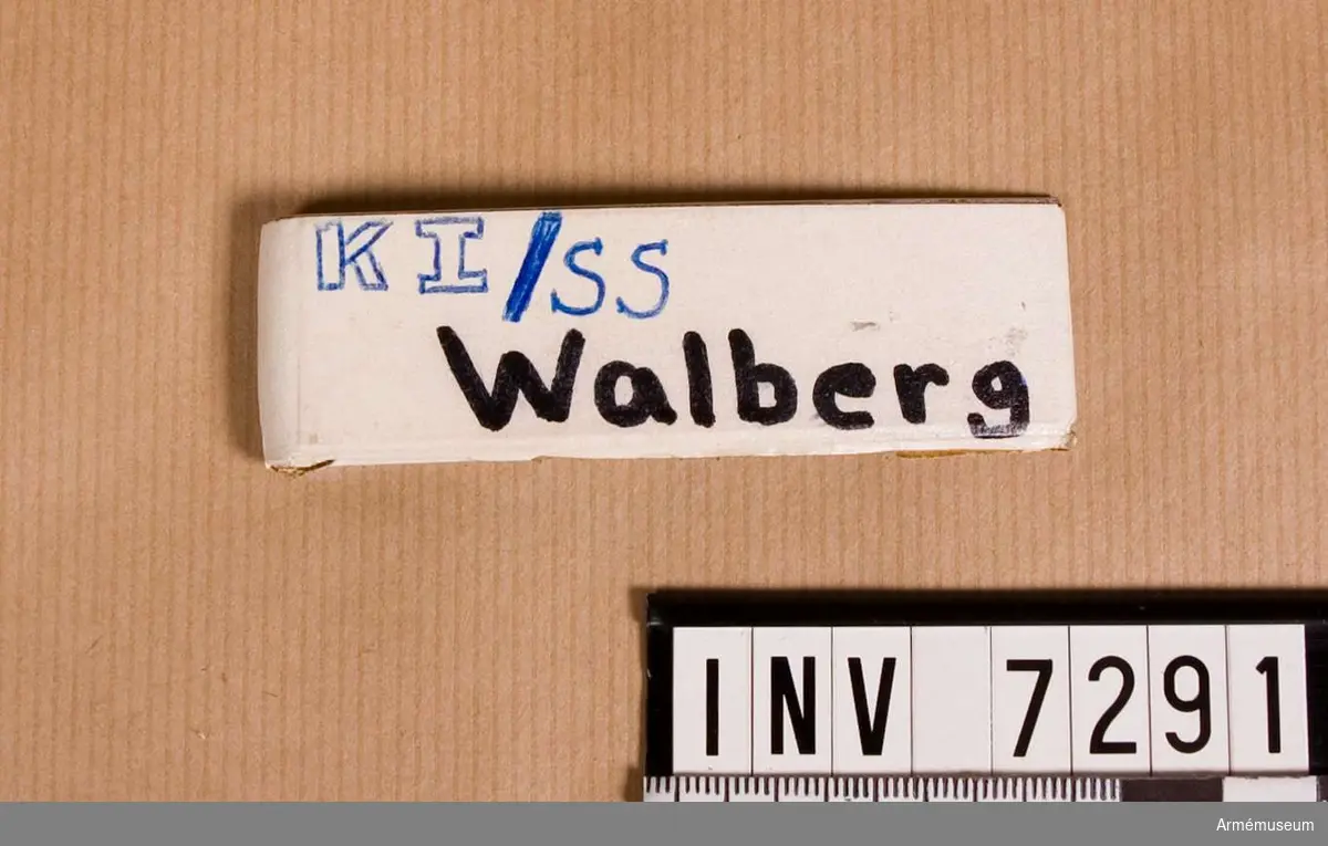 Utförd i aluminium med klämma. På framsidan påsatt en remsa av vit tape. På tapen är skrivet med svart spritpenna: Walberg. Däröver är textat med bläck: KI/SS. 