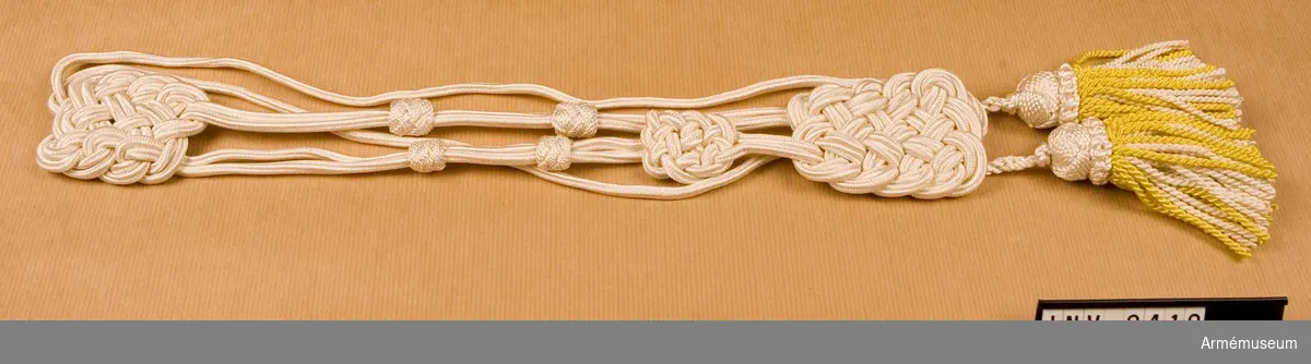 Av vit silkessnodd och gult silke i tofsens yttre skikt, med vitt silke i dess inre.
Källa: UNI:A 1977 mom 501 och 502. Tillhör utrustning för  musikavdelningar och vakter. 
