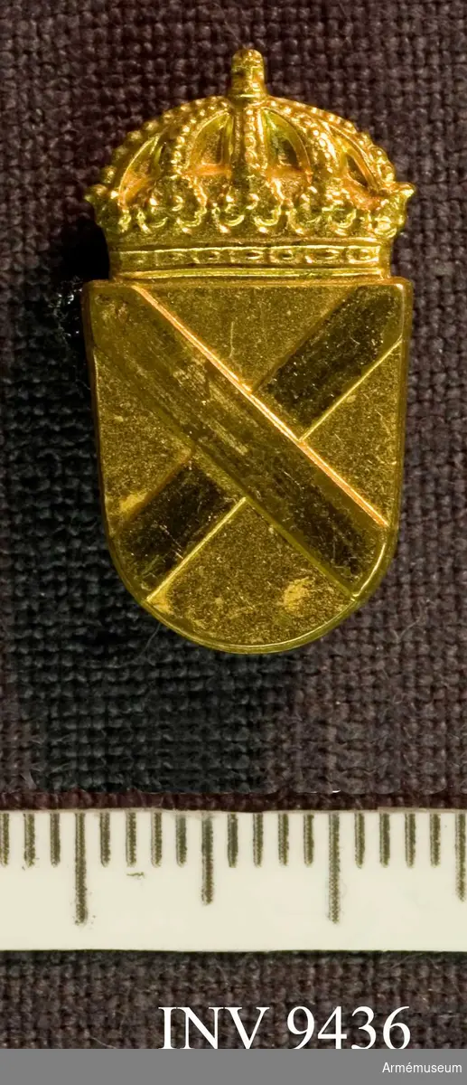 Samhörande nr är 9421 - 9438.
Civilförsvarsmärke i guld, SLK.
Ett förgyllt märke, troligen tillverkat hos Sporrong. Ingen märkning finns synlig. Framsidan visar två korslagda värjspetsar vilande på en sköld, krönt av en kunglig krona.