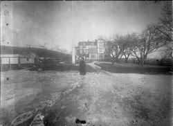 Kvinne stående foran flaggstang i hage, stort trehus i bakgr