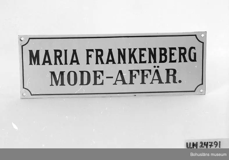 Emaljerad. Vit bakgrund, blå text: "MARIA FRANKENBERG MODE-AFFÄR." Blå ram runt texten.

Modisten Maria Frankenberg (1858-1938) startade den första modeaffären hösten 1886 på Asplundsgatan. Därefter innehade hon modehandel på Norra Drottninggatan 1, Uddevalla, Maria Frankenberg Mode-affär. Affären övertogs av bl.a. Sigrid Hansson efter Frankenbergs död och hette därefter "Maria Frankenbergs Eftr. Modeaffär Uddevalla". 
Se även UM022807:1-2; skyltar från samma modeaffär.