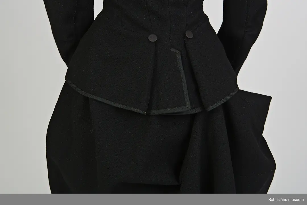 Svart lång kjol sydd i kypertvävt ylletyg.
Del av riddräkt UM62.04.001 - UM62.04.007
Kjolen är oregelbundet formad. Längsta delen på höger baksida är längst. Vid användning häktas denna upp med hjälp av en hake och en sydd hank (skadad).
Enkel bandkantad linning med hyske och hake samt snedställd öppning som knäppes av fyra tygklädda knappar. Samma typ som i jackan. 
Fållens uppvikta kant är sicksackklippt samt uppsydd med maskinsöm.
Mer information se UM62.04.001