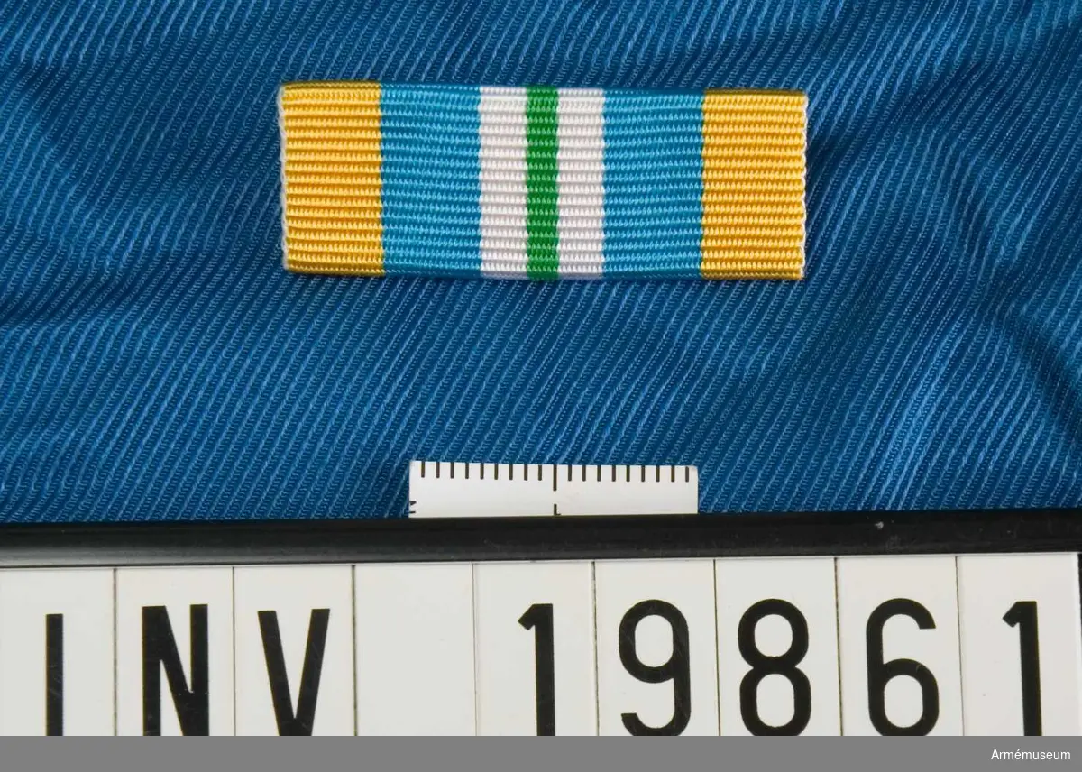 Blått band med ett grönt streck på mitten åtföljd på vardera sidan av en vit rand samt med breda gula kanter. Släpspännet förvaras i ask tillsammans med en medalj.