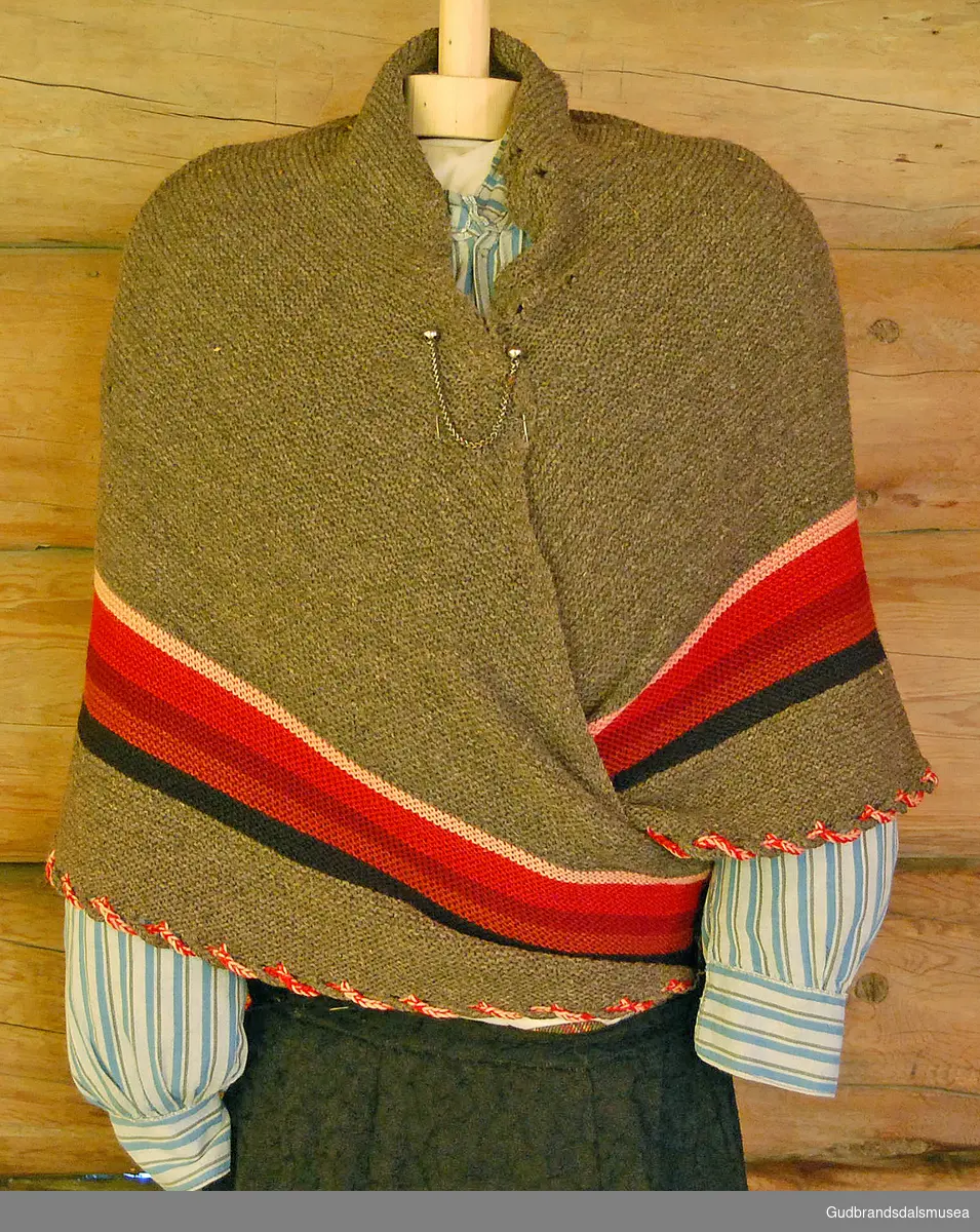 Bondetørkle i strikket ull, med striper i ulike farger, knyttet bånd i rødt og hvitt rundt halve tørklet, heklet snor. To nåler i metall med lenker til å sette sammen/feste tørklet ved bruk. 