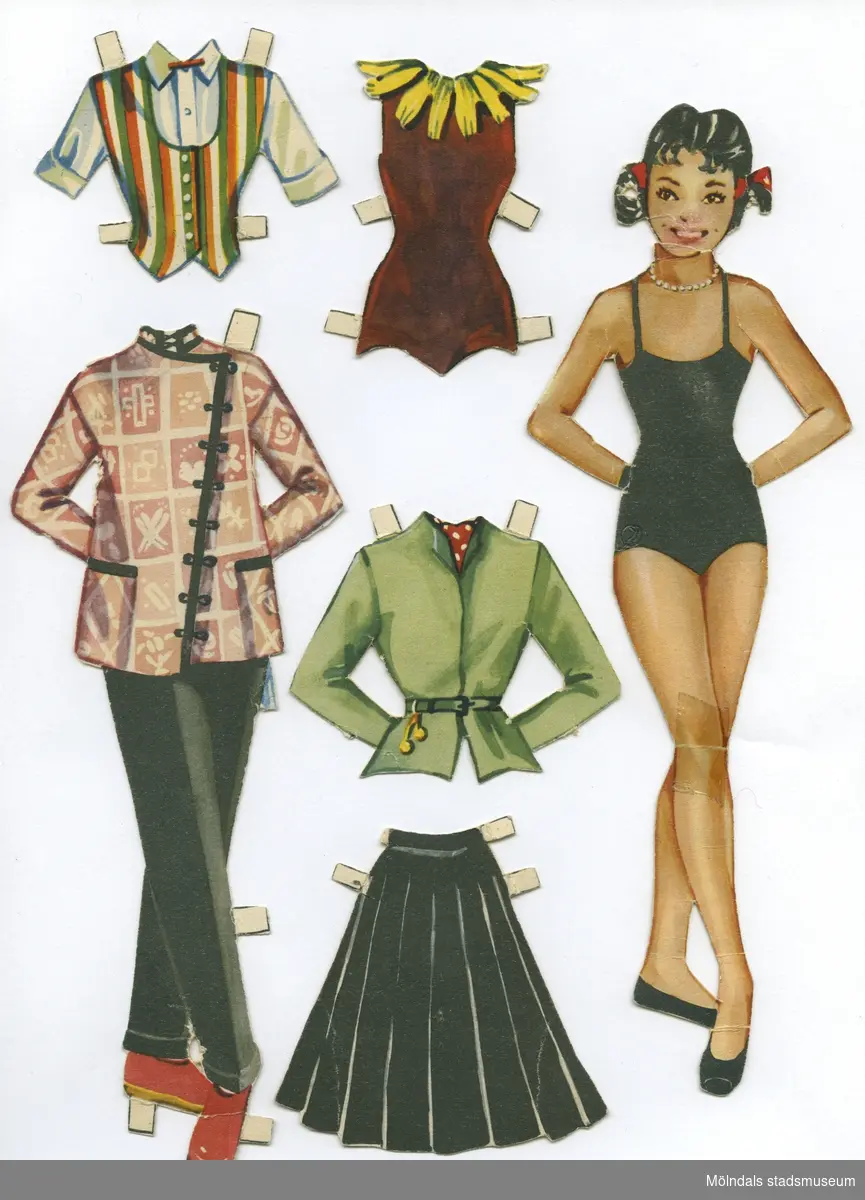 Pappdocka med kläder från 1950-talet. Docka och kläder är märkta "Margot" på baksidan - dockans namn.Dockan föreställer en ung kvinna med svart hår i tofsar, iklädd svart baddräkt och lågskor. Garderoben består av set med byxor och orientalisk jacka, toppar, kjolar, ballerinadräkt, östeuropeisk (?) folkdräkt, samt skor med snörning upp på smalbenet. Docka och kläder förvaras i ett brunt kuvert, märkt "Margot", men är ursprungligen ett utgivarekorsband från Handelsnytt, Malmö.