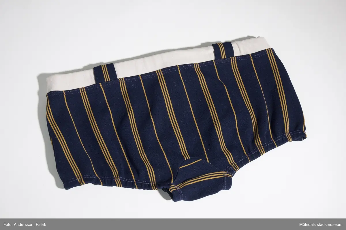 Mörkblåa badbyxor med gula ränder och ett vitt band i midjan. Dessa användes av givarens man Alf (f. 1943) under 1960-talet.MåttLängd i sidan: 165 mm, Midjebredd: 330 mm