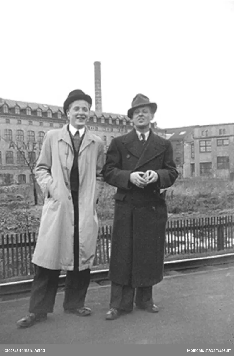 Charlie Jinnerstedt och vännen Helmer Garthman står på en väg, uppklädda i hatt och rock. I bakgrunden syns Almedals fabriker. Almedal (Göteborg), 1930.