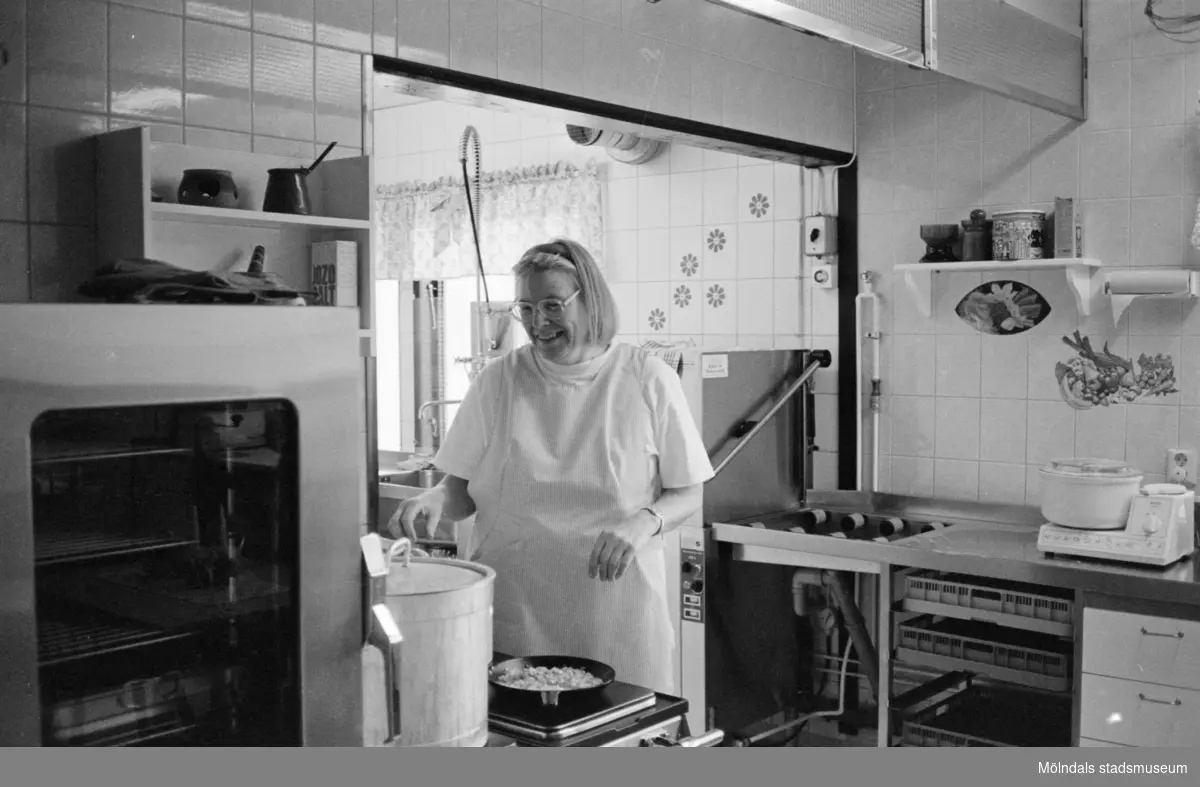 En blond kvinna, med glasögon och förkläde, står i Katrinebergs daghems kök och lagar mat vid spisen. Runt omkring finns ugn, diskbänk, skåp och köksmaskiner.