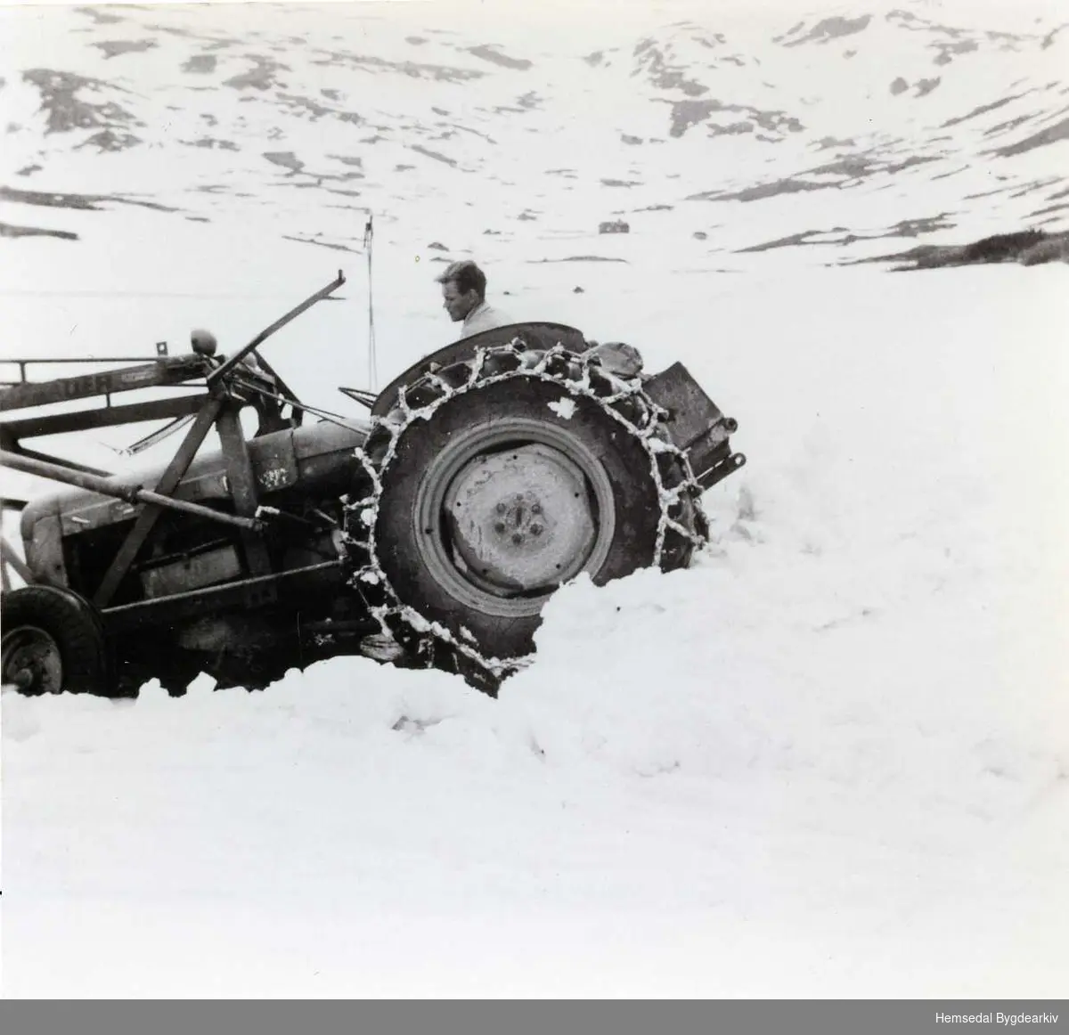 Innkøyring av bulldosar, traktor og utstyr i 1959 til arbeid med demminga av Dyrja på Feten i Hemsedal.
Transporten er her komen noko beint ved oset i nordenden av Svartevatnet.
Eirik Snerte køyrer traktoren.
