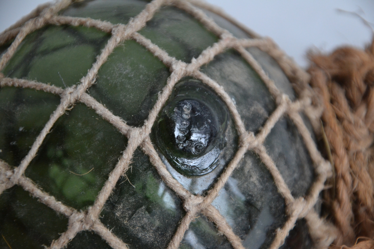 En grønn glasskule med nett av bomullstråd festet rundt seg. En hempe av kokostau er festet i den ene enden. På glasskula er merket et symbol med seks rundinger vannrett og tre rundinger loddrett. Nettet er tvinnet på plass, ikke knytt.