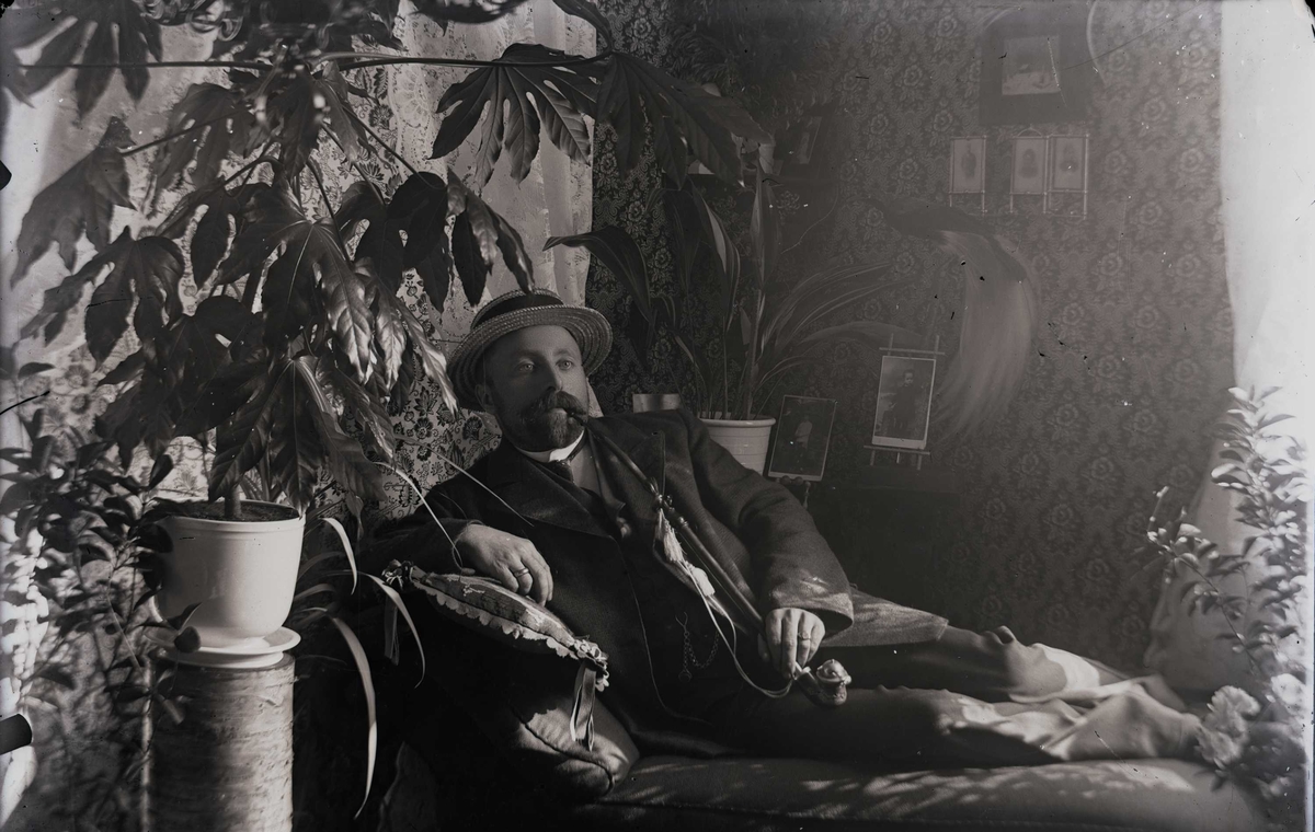 Portrett av mann som ligger på en divan i en pent innredet stue, mens han røker på en langpipe.