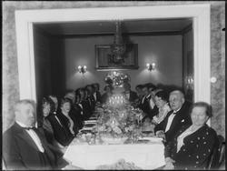 Stueinteriør med kvinner og menn, festpyntet. Ukjent.