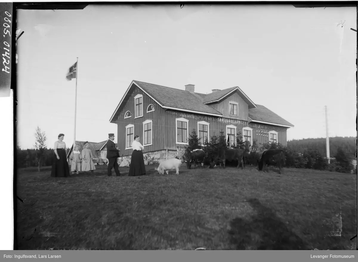 Skolebygning med to kvinner, en mann, to barn og husdyr i forgrunnen.