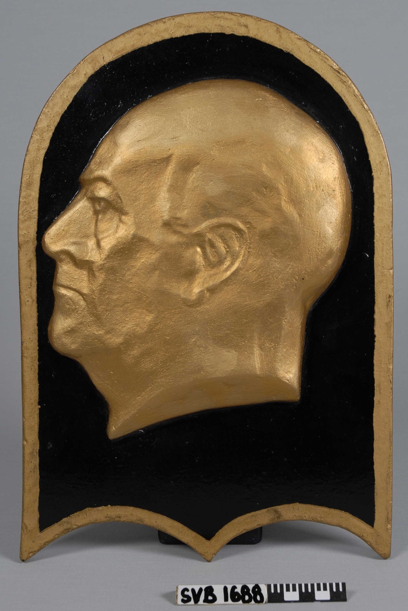 Plakett i sort og gull. Relieff av Kong Olav V i profil.