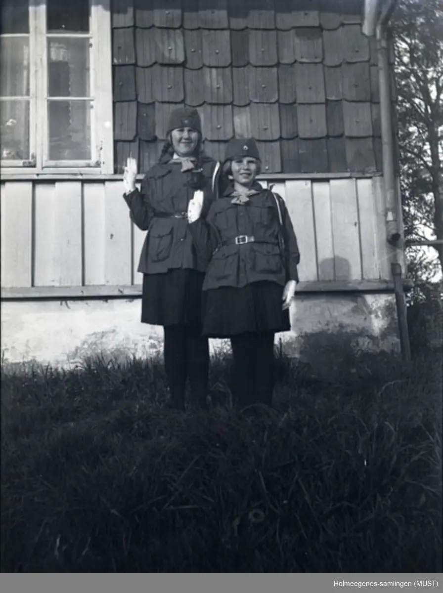 Gunhild og Karen Pedersen i speideruniformer foran huset på Holmeegenes. Begge gjør speiderhilsen til fotografen.