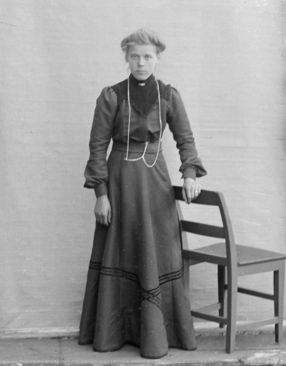 Ung kvinne kledd i mørk kjole, avbildet stående ved stol, foran lerret på husvegg