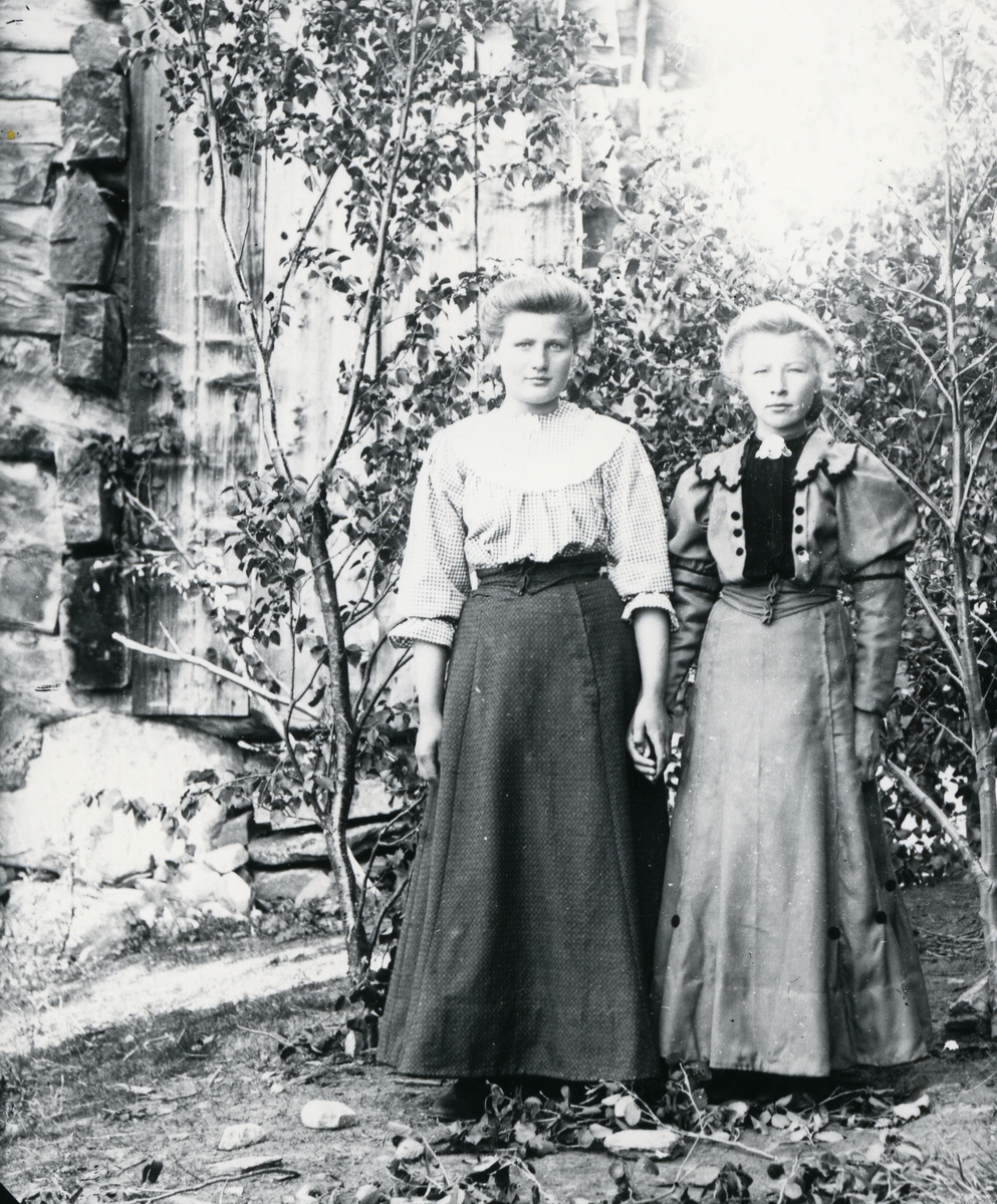 To kvinner i helfigur som holder hverandre i hendene. Stående foran bjørkekvister og tømmervegg