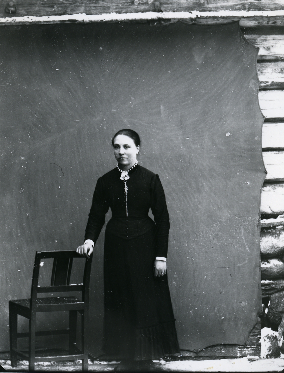 Kvinne i helfigur, stående ved stol, foran lerret