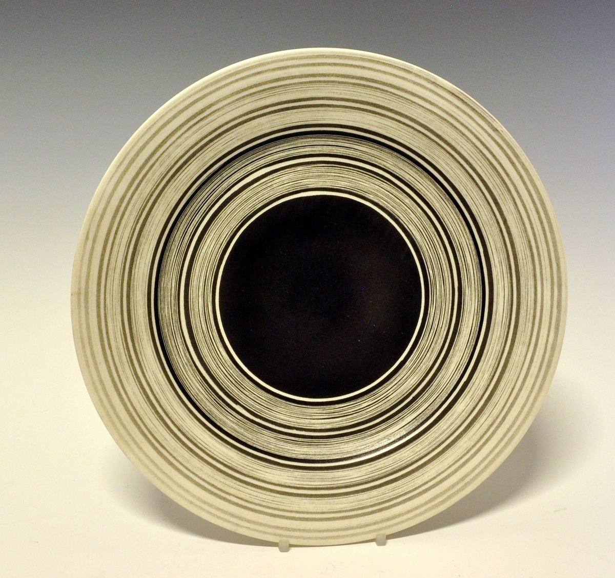 Flat tallerken av porselen. Hvit glasur. Midterste del av speilen er sortmalt, ut fra denne sorte og hvite striper i ulike bredder.