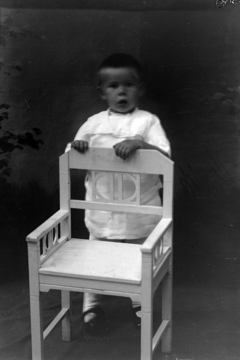 Studioportrett av et lite barn som står bak en stol.