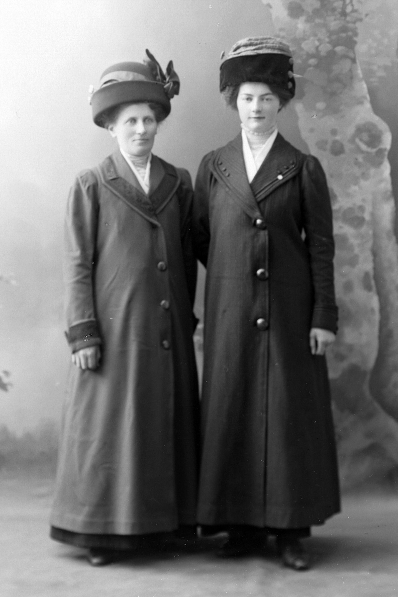 Studioportrett av to kvinner i frakk med hatter på hodet.