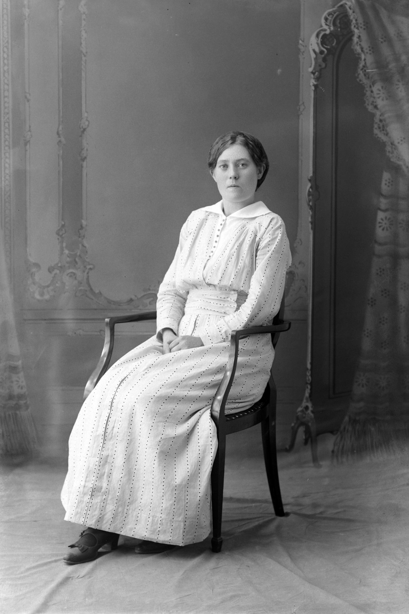 Studioportrett av en sittende kvinne i hvit kjole.