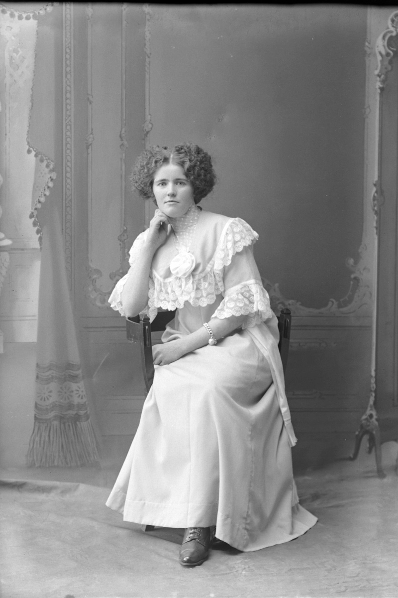 Studioportrett av en sittende kvinne med lys kjole.