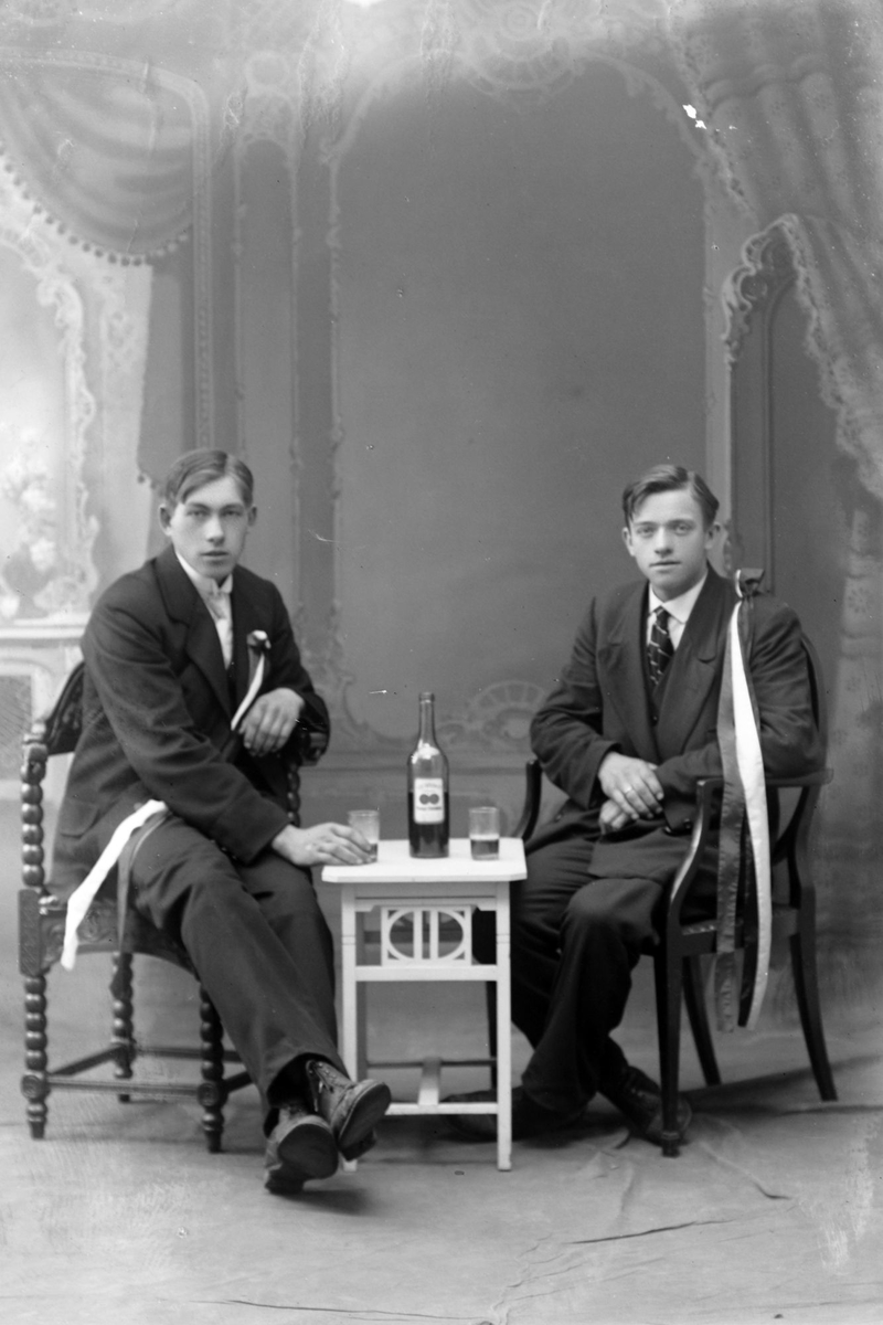 Studioportrett av to menn med store sløyfer. De sitter ved et bord og deler en flaske.