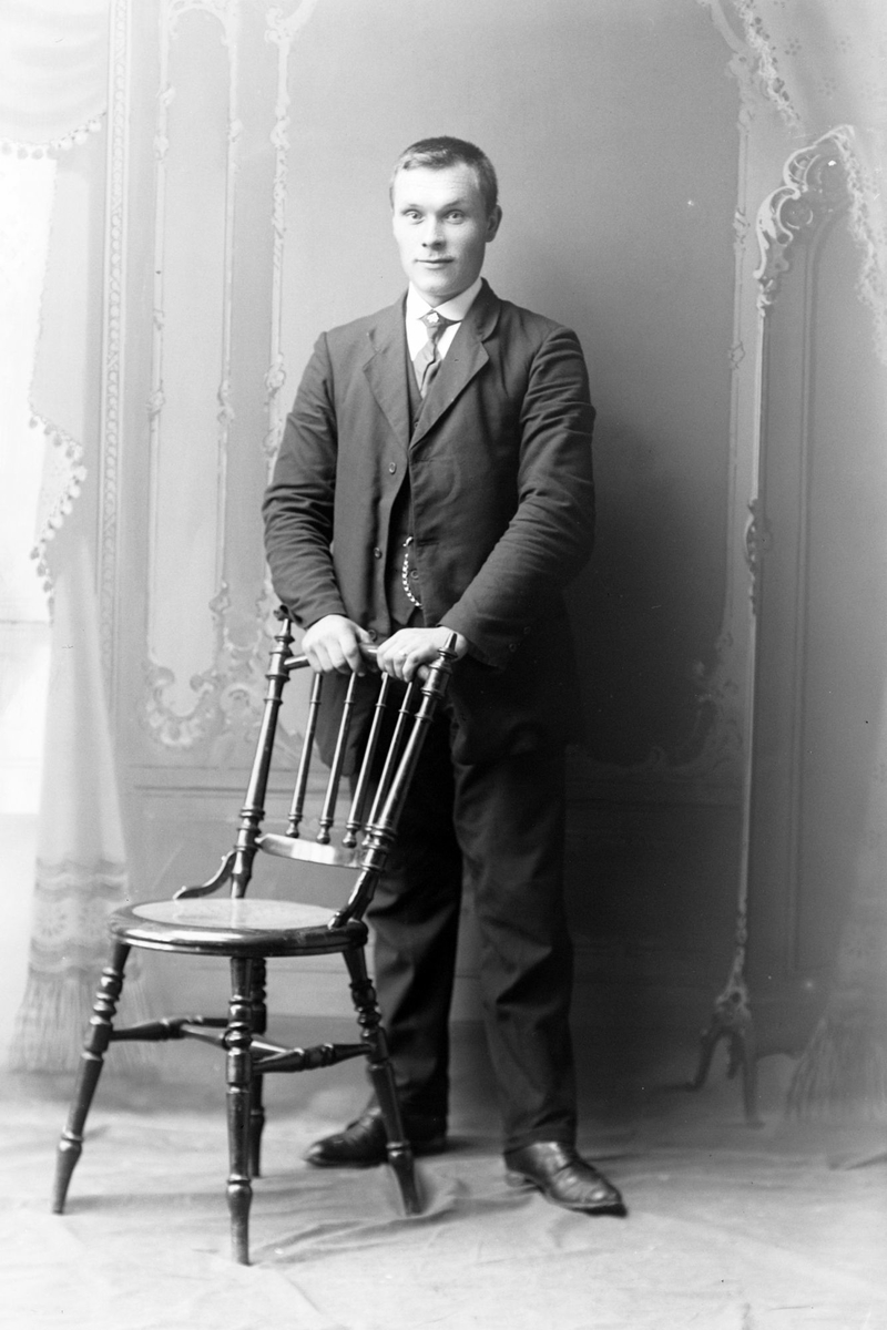Studioportrett av en mann stående bak en stol.
